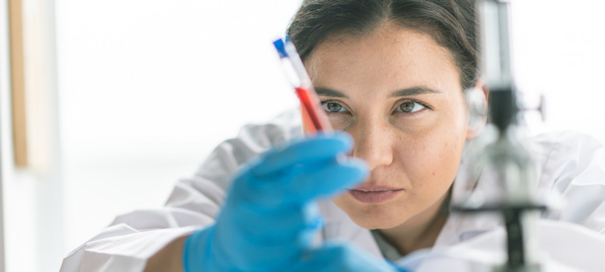 <p>A female laboratory technician examines blood vial sample. Medical gloves and a lab coat are worn for protection. Focus is on the laboratory technician.</p>